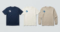 The sand long sleeve tee shown alongside the navy athletic tee and off-white short sleeve, each carrying the Cross at the right chest.