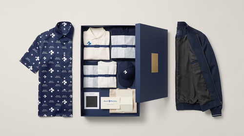 The Founder Kit, Navy: the open presentation box with the standard garments folded and wrapped inside, the Silk Beach Shirt and the Basalt-Lined Bomber displayed individually beside it. Design concept rendering.