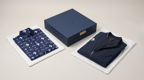 The closed navy presentation box with the engraved edition plate, the Silk Beach Shirt and the Basalt-Lined Bomber displayed individually on their dust bags either side. Design concept rendering.